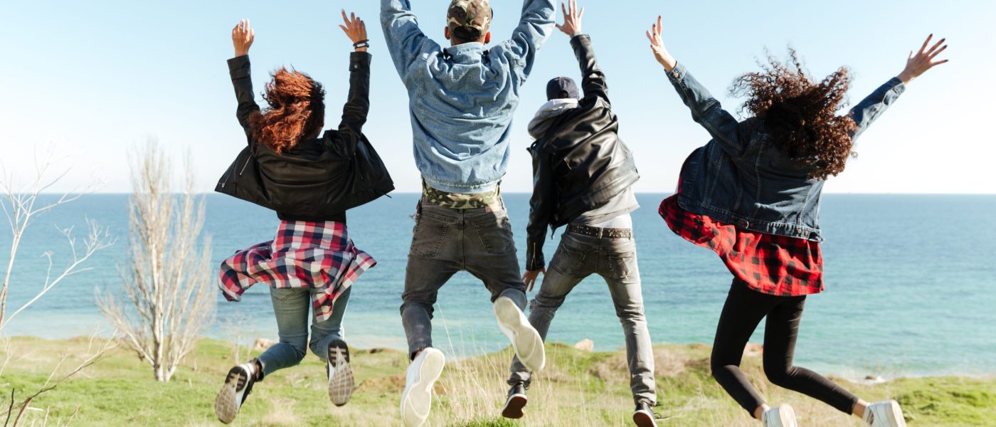 Back view picture of a group of friends jumping outdoors near beach with raised hands.
